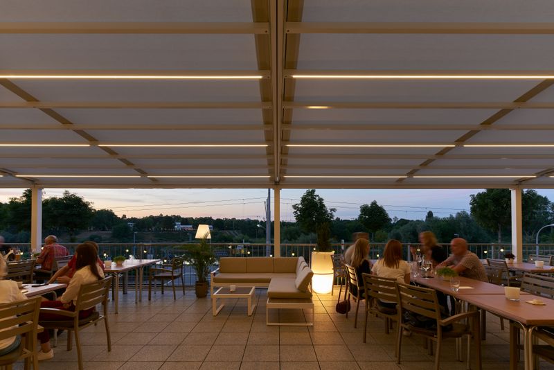 Reference image of a pergola stretch with cream fabric cover and white frame over the outdoor area of the "bootshaus" in Mannheim at dusk. LED-Lines can be seen and provide cozy light.